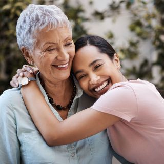 A photo of two happy women. The younger one has her arm draped around the neck of the older one, embracing her.