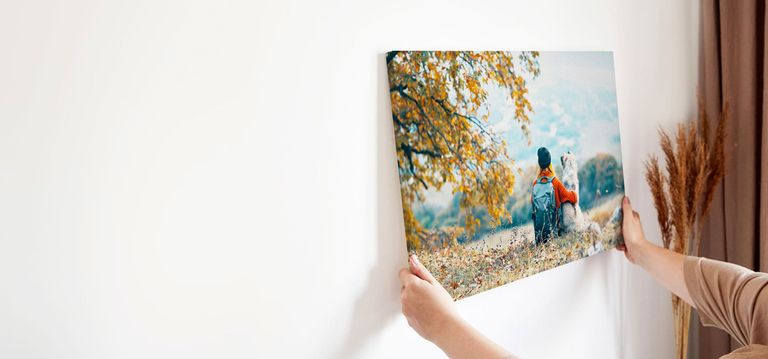 A person hanging a canvas print on a white wall. The canvas features a serene landscape of a dog and their owner.