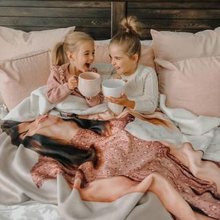 Two girls on a bed wrapped in a custom photo blanket, smiling and holding mugs.