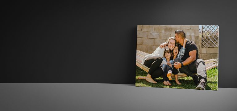 Canvas print featuring a joyful family of four sitting on a hammock outdoors, with the father kissing the mother on the forehead while their children smile and embrace each other.